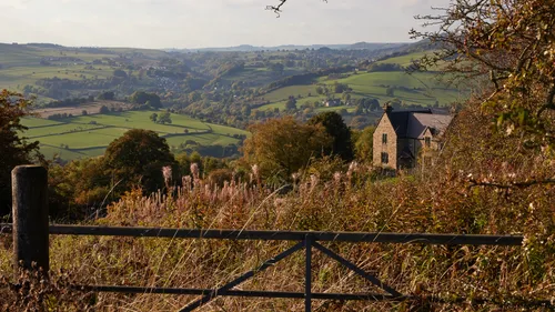 This landscape photograph was taken in the afternoon during autumn and shows a rural Peak District valley in Derbyshire, United Kingdom. The main subject of the image is a metal gate overlooking a sweeping vista filled with autumnal nature, including tall grasses and shrubs in the foreground and rolling, green fields beyond. A traditional stone house is partially visible among the trees to the right, while the view stretches across the valley to distant countryside dotted with woodlands and small settlements. Curbar Edge, a prominent gritstone escarpment and well-known landmark in the Peak District, forms part of the undulating hills in the background. The image captures the rural charm and natural landscape of the Peak District during the autumn season.